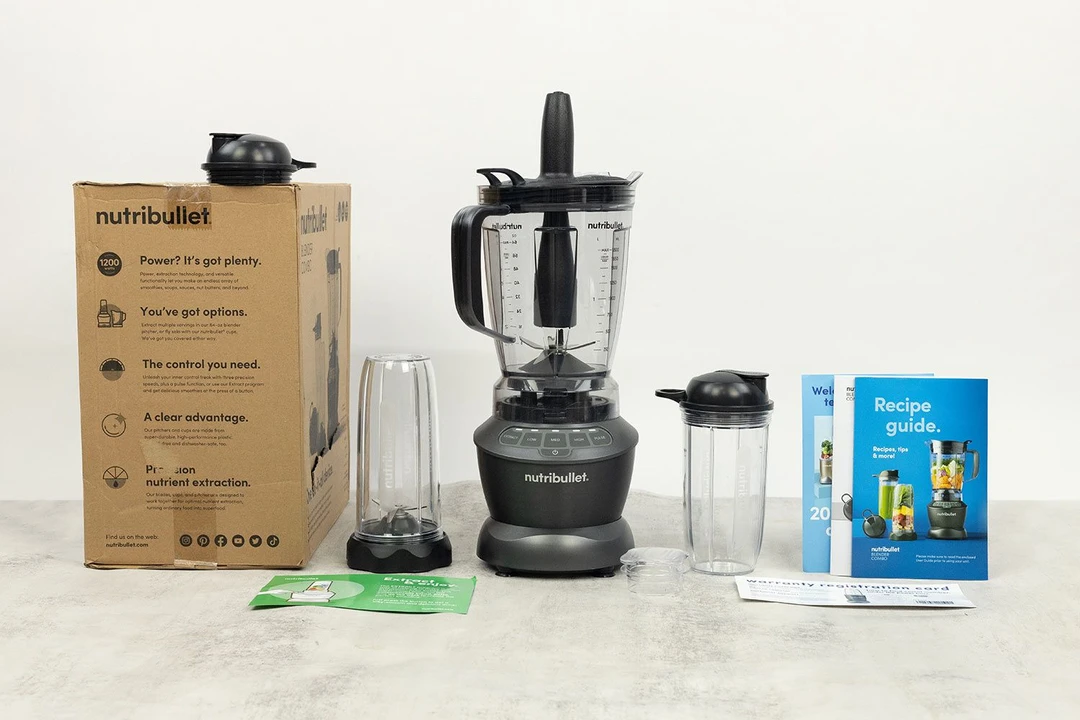 The NutriBullet ZNBF30500Z Blender Combo stands on a table, with a recipe booklet, a to-go cup with lid, an extra blending cup with blade assembly attached, a lid, and a carton box by its sides.