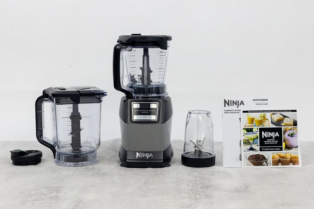 The Ninja AMZ493BRN blender stands on a table, with a 72-oz blending container, 18-0z blending cup, lid, user manual, and a carton box by its sides.
