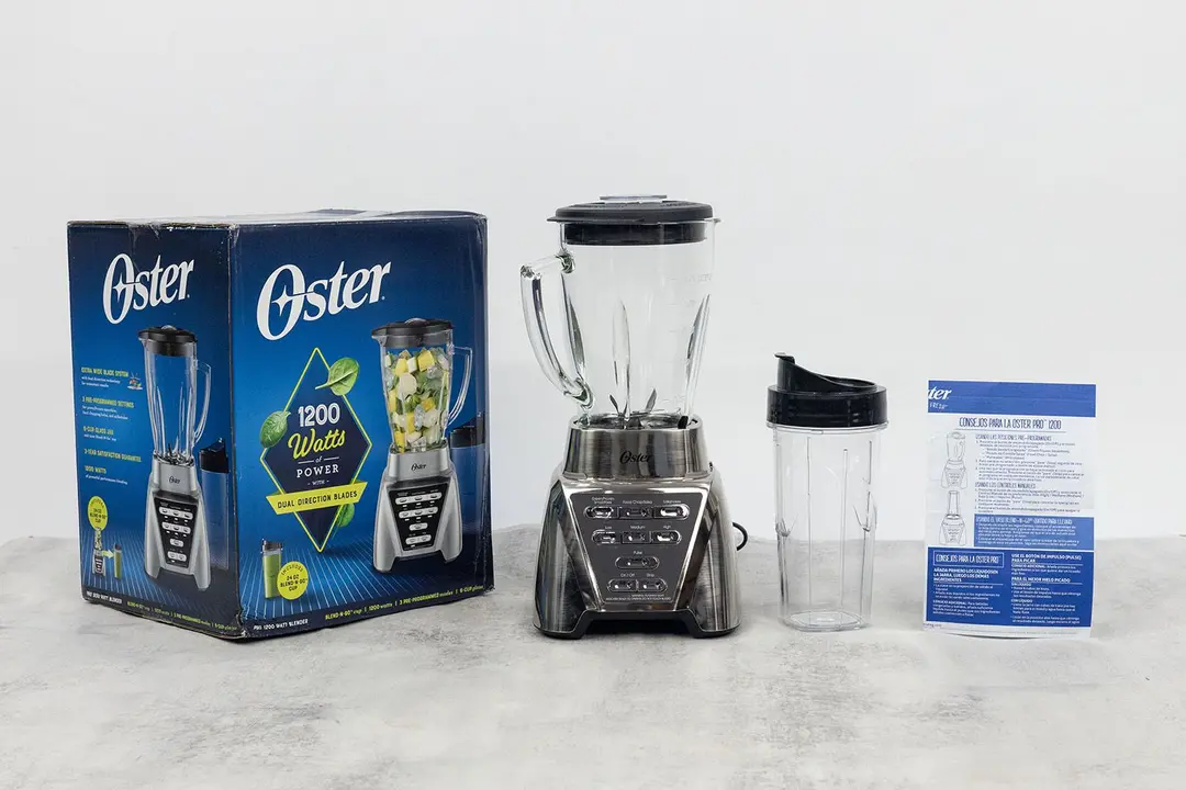 The Oster Pro blender stands on a table, with a recipe booklet, a to-go cup with lid, and a carton box by its sides.