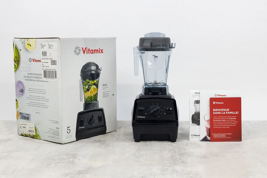 The Vitamix E310 blender stands on a table, with a recipe booklet, a lid plug, and a user manual by its sides.