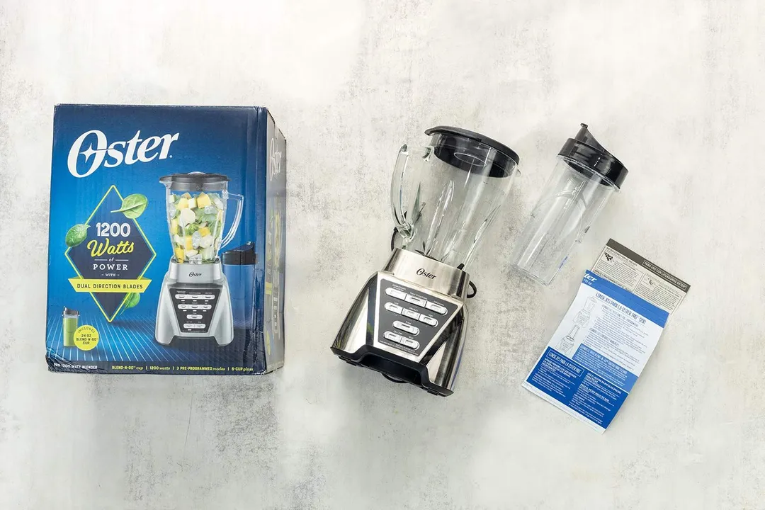 A display of the Oster Pro blender and its accessories, including the user manual, 24 oz cup with lid, and carton box, on a table.