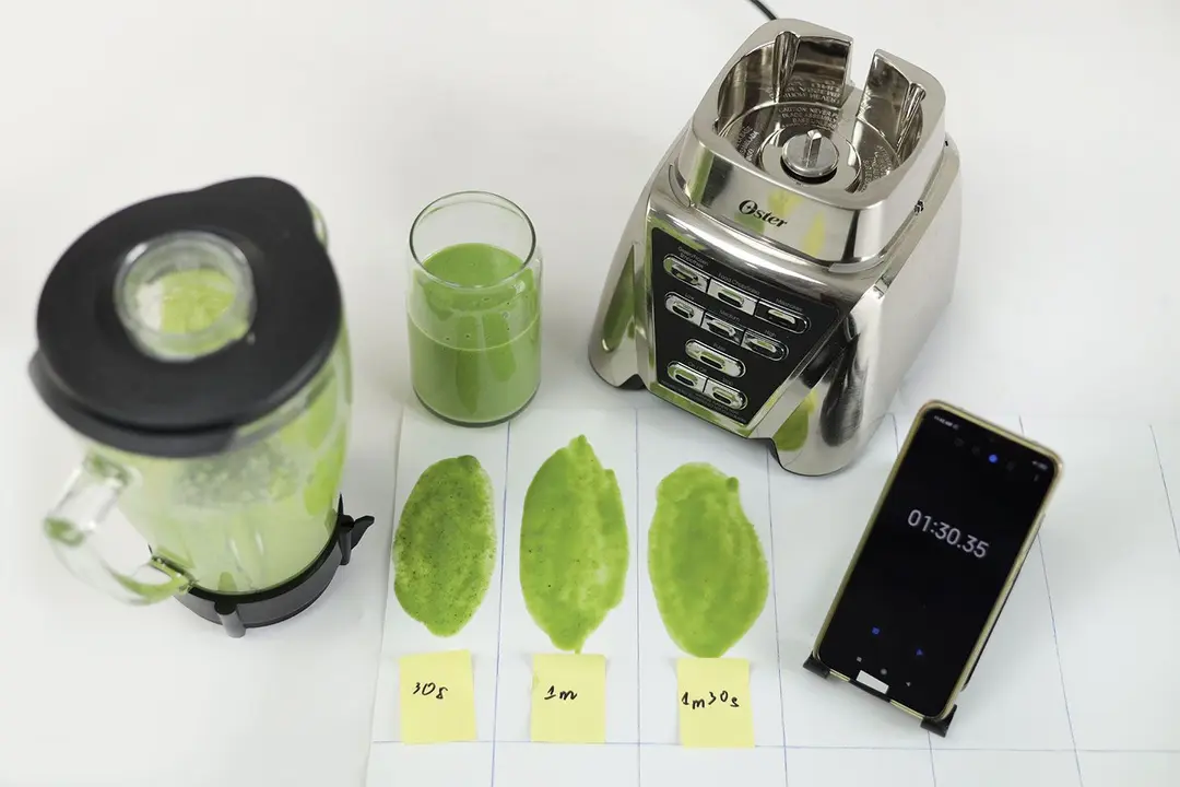 The Oster Pro motor base and its container stand on a table, accompanied by a glass filled with a green smoothie. Next to it, a white paper features two distinct smoothie samples while a smartphone showcases the blending time of 1 minute and 30 seconds