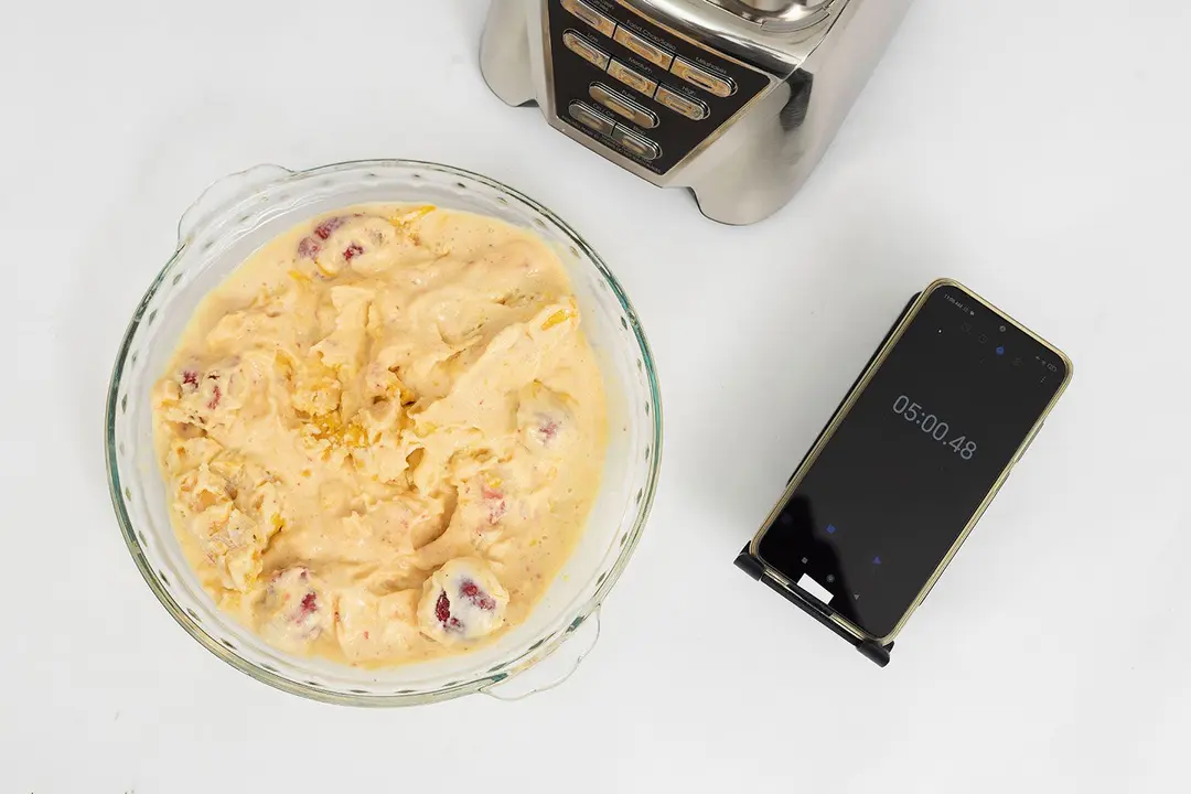 The Oster Pro motor base stands beside a glass bowl which contains a portion of the smoothie it produced. Next to it, a smartphone displays a blending time of 5 minutes.