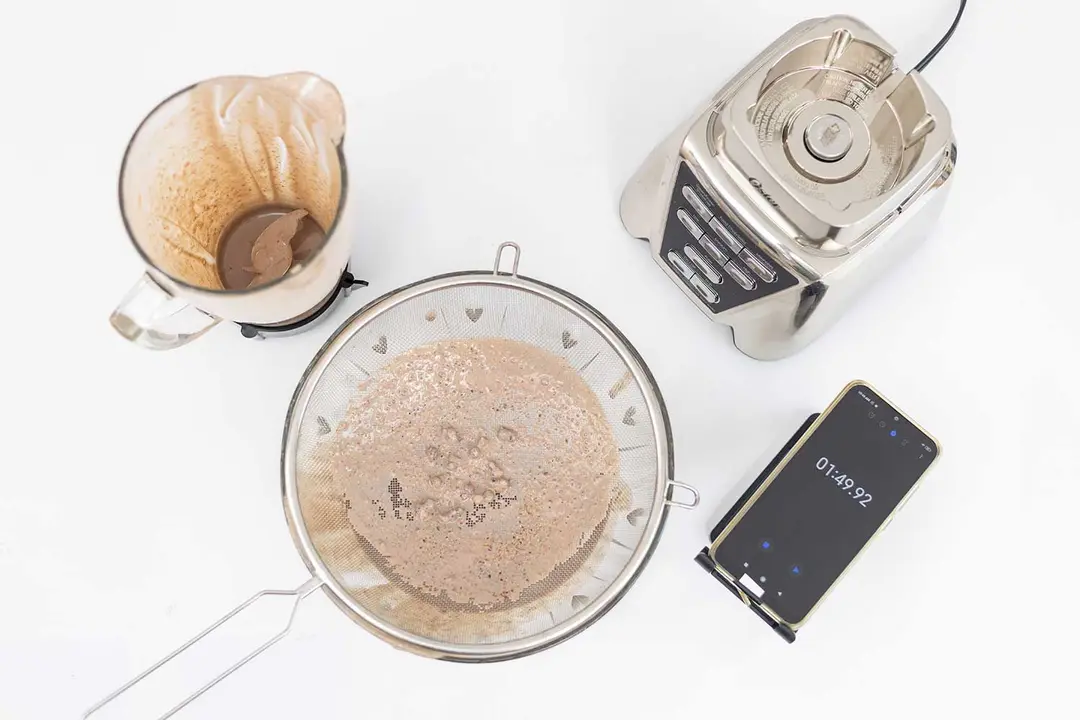 The Oster Pro  motor base stands beside the container. Next to it, a protein shake has been strained through a metal mesh strainer while a smartphone displays a blending time of 1 minute 49 seconds. 