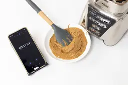 The Oster Pro is beside a white plate containing almond butter with a spatula and a smartphone revealing a blending time of 6 minutes.