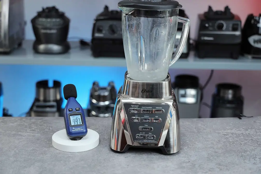 The Oster Pro blender is on a table, accompanied by a sound level meter displaying its noise level nearby. In the background, other blenders that we tested sit on a nearby shelf.