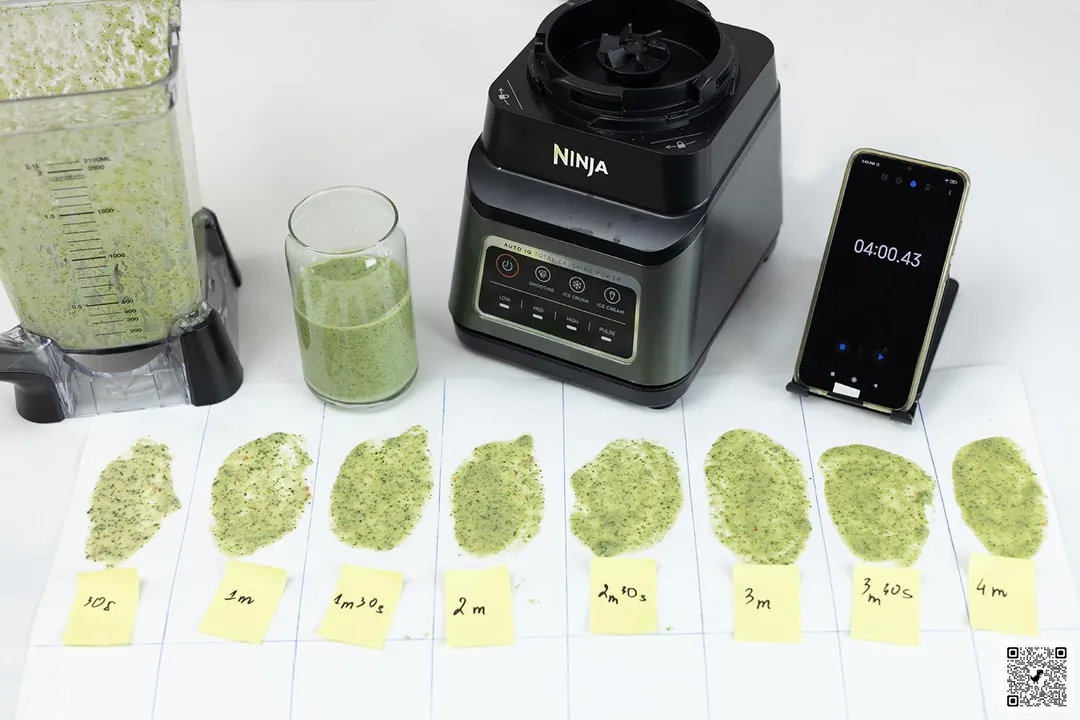The Ninja BN701 Professional Plus blender’s motor base and its container stand on a table, accompanied by a glass filled with a green smoothie. Next to it, a white paper features three distinct smoothie samples while a smartphone showcases the blending time of 4 minutes.