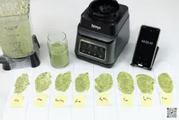 The Ninja BN701 Professional Plus blender’s motor base and its container stand on a table, accompanied by a glass filled with a green smoothie. Next to it, a white paper features three distinct smoothie samples while a smartphone showcases the blending time of 4 minutes.