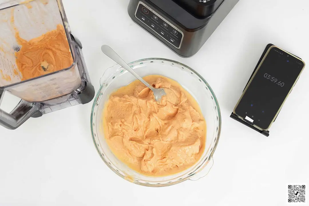 The Ninja BN701 Professional Plus Blender motor base stands beside a glass bowl which contains a portion of the smoothie it produced. Next to it, a smartphone displays a blending time of 3 minutes and 59 seconds.