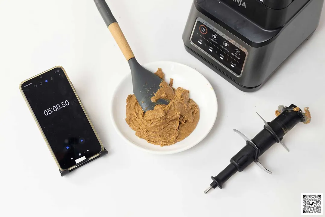 The Ninja BN701 Professional Plus is beside a white plate containing almond butter with a spatula, blend assembly, and a smartphone revealing a blending time of 5 minutes.