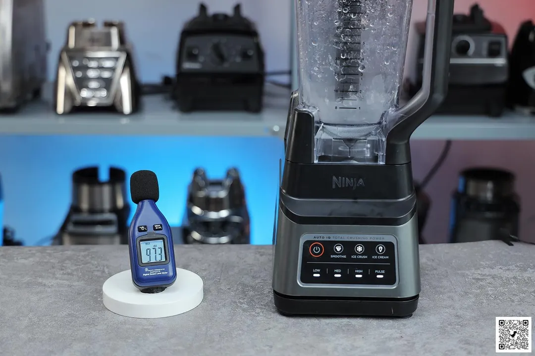 The Ninja BN701 Professional Plus blender is on a table, accompanied by a sound level meter displaying its recorded noise level. In the background, other blenders that we tested sit on a nearby shelf.
