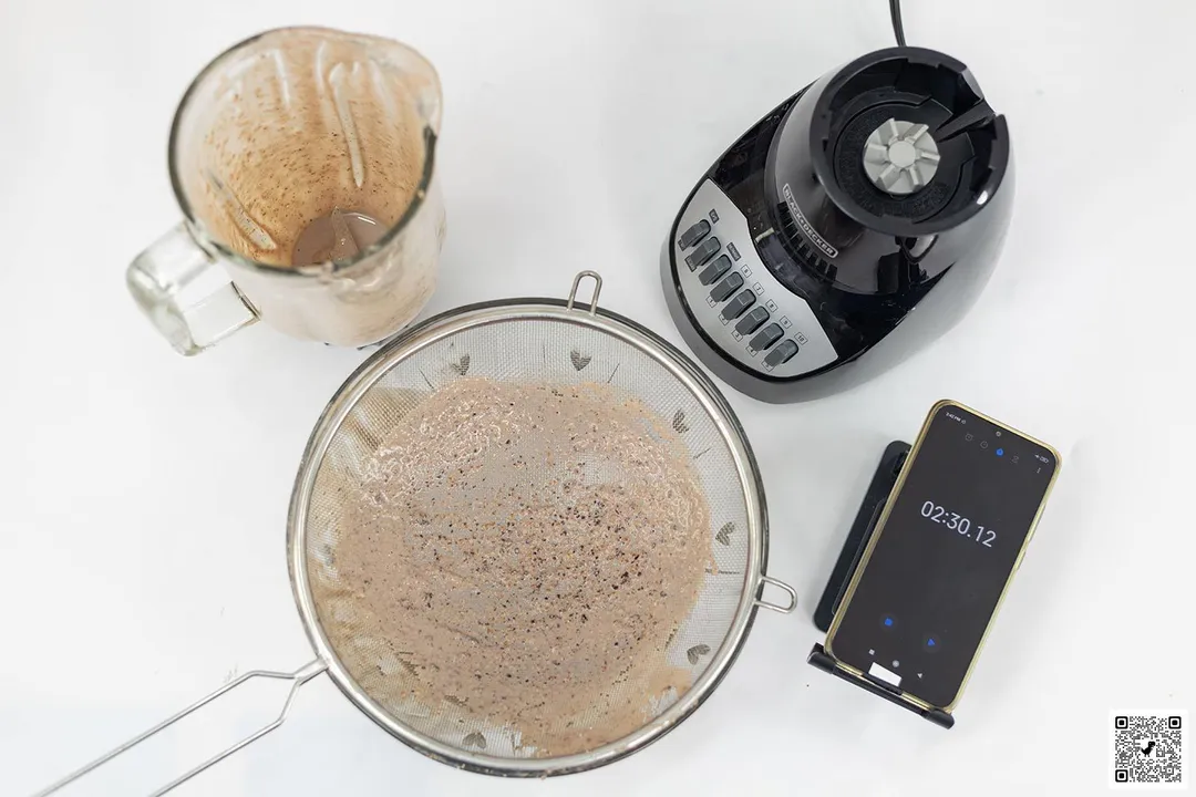 The Black+Decker Crush Master motor base stands beside the container. Next to it, a protein shake has been strained through a metal mesh strainer while a smartphone displays a blending time of 2 minutes 30 seconds.