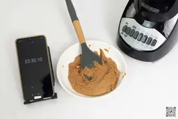 The Black+Decker Crush Master is beside a white plate containing almond butter with a spatula and a smartphone revealing a blending time of 3 minutes and a half.
