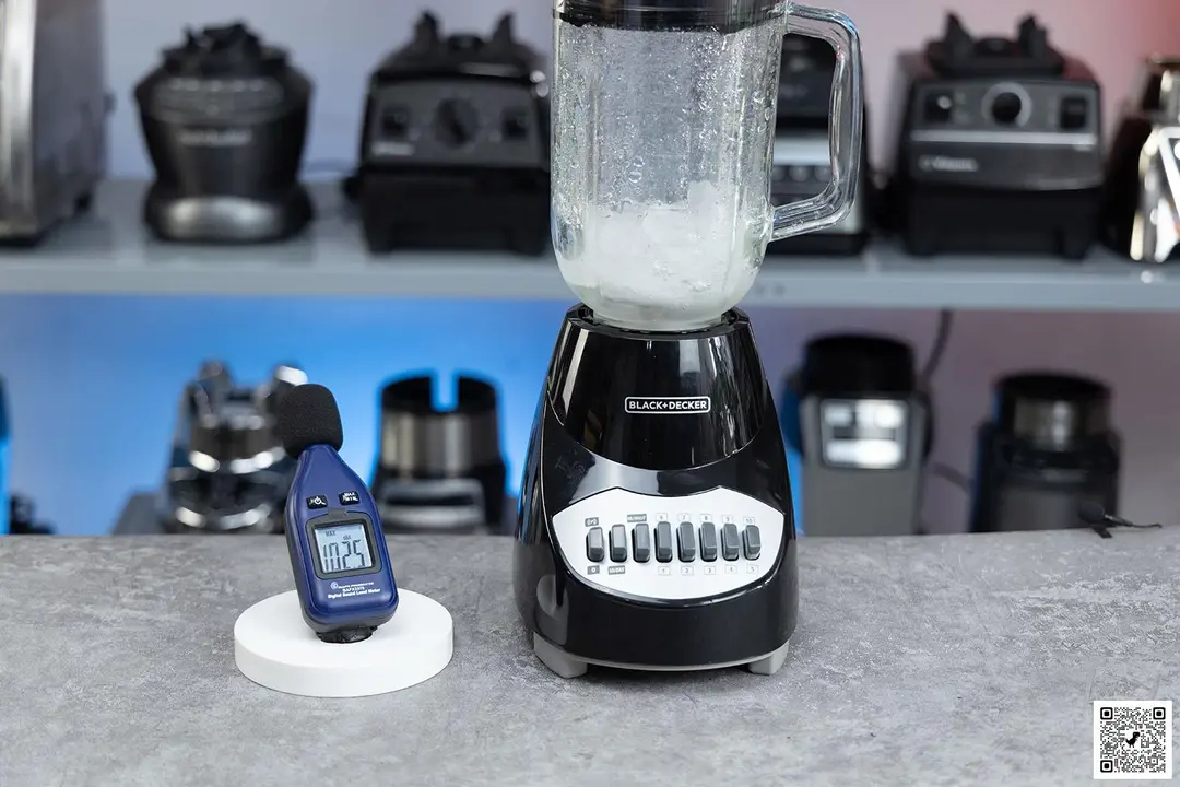 The Black+Decker blender is on a table, accompanied by a sound level meter displaying its noise level nearby. In the background, other blenders that we tested sit on a nearby shelf.