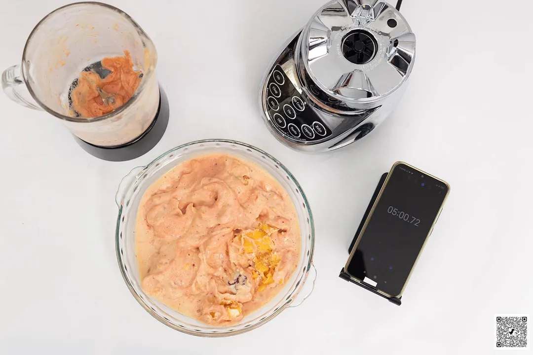 The Cuisinart SmartPower motor base stands beside a glass bowl which contains a portion of the smoothie it produced. Next to it, a smartphone displays a blending time of 5 minutes.