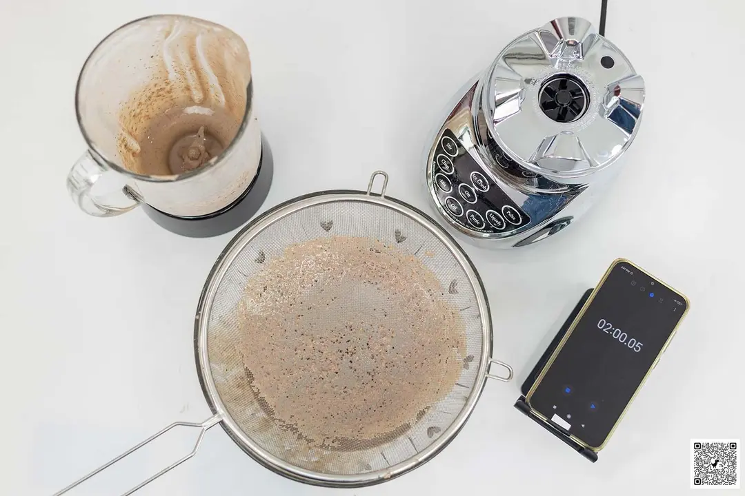 The Cuisinart SPB-7CH SmartPower motor base stands beside the container. Next to it, a protein shake has been strained through a metal mesh strainer while a smartphone displays a blending time of 2 minutes.
