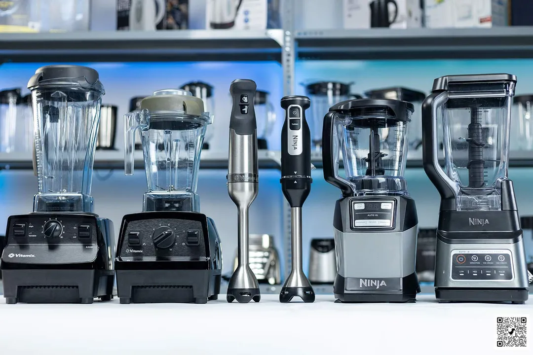 Vitamix E310, Vitamix 5200, Vitamix immersion blender, Ninja immersion blender, Ninja AMZ.. and Ninja BN701 neatly arrayed on a countertop. Various other blender models display on the shelves in the backdrop.