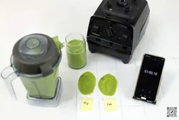 The Vitamix E310 motor base and its container stand on a table, accompanied by a glass filled with a green smoothie. Next to it are two distinct smoothie samples spread on white paper while a smartphone showcases the blending time of 1 minute.