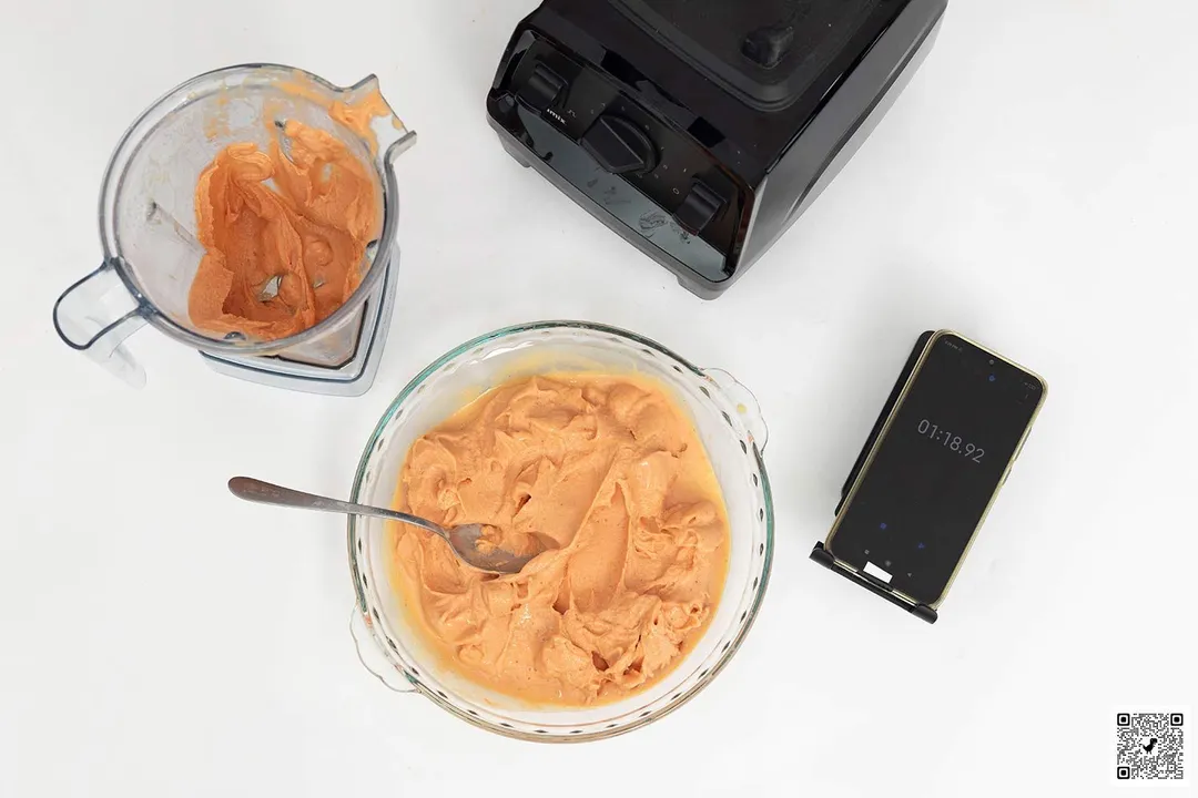 The Vitamix E310 motor base stands beside the container, which holds a portion of the smoothie it produced. Next to it, a glass bowl contains the remaining smoothie while a smartphone displays a blending time of 1 minute and 20 seconds.