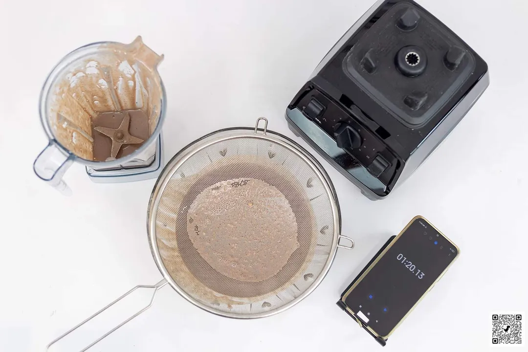 The Vitamix E310 motor base stands beside the container. Next to it, a metal mesh strainer filtered its protein shake while a smartphone displayed a blending time of 1 minute 20 seconds.