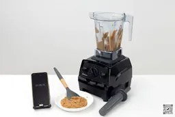 The Vitamix E310 is beside a white plate containing almond butter and a spatula, and a smartphone revealing a blending time of 1 minute and a haft.
