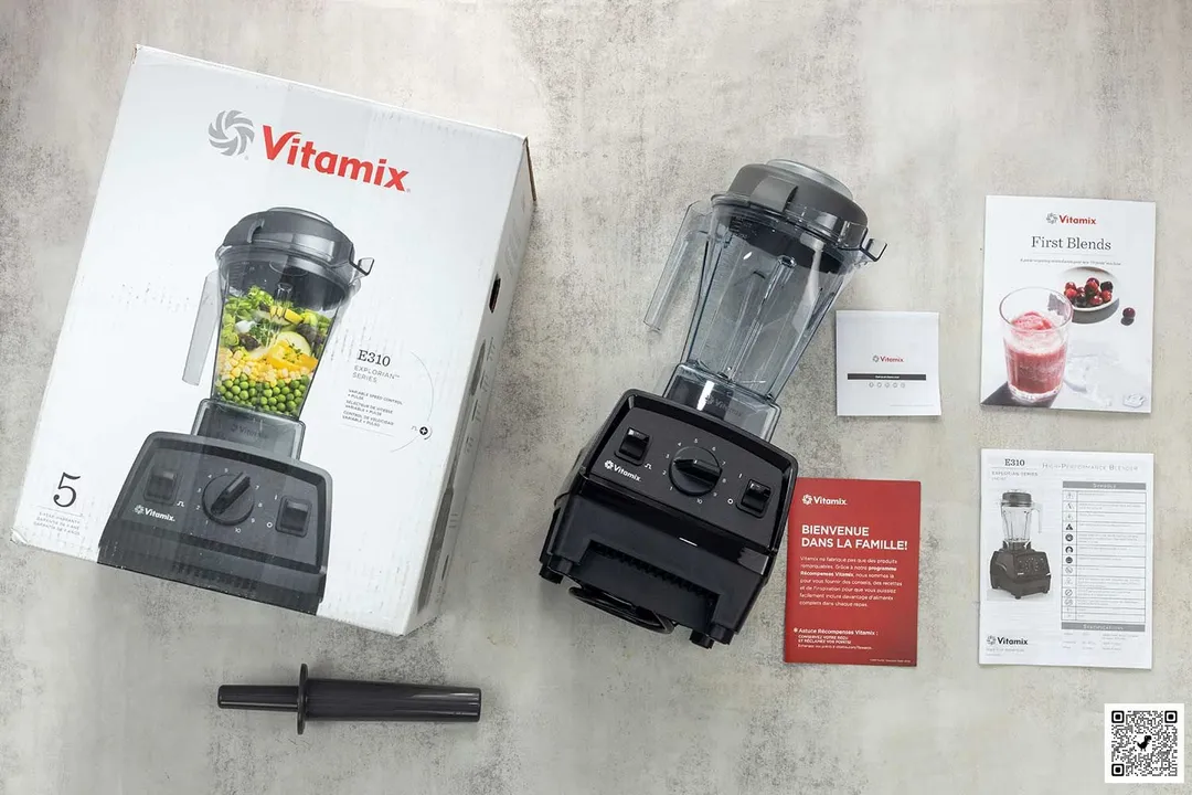 A display of the Vitamix E310 blender and its accessories, including the tamper, carton box, recipe booklet, and user manual, on a table.