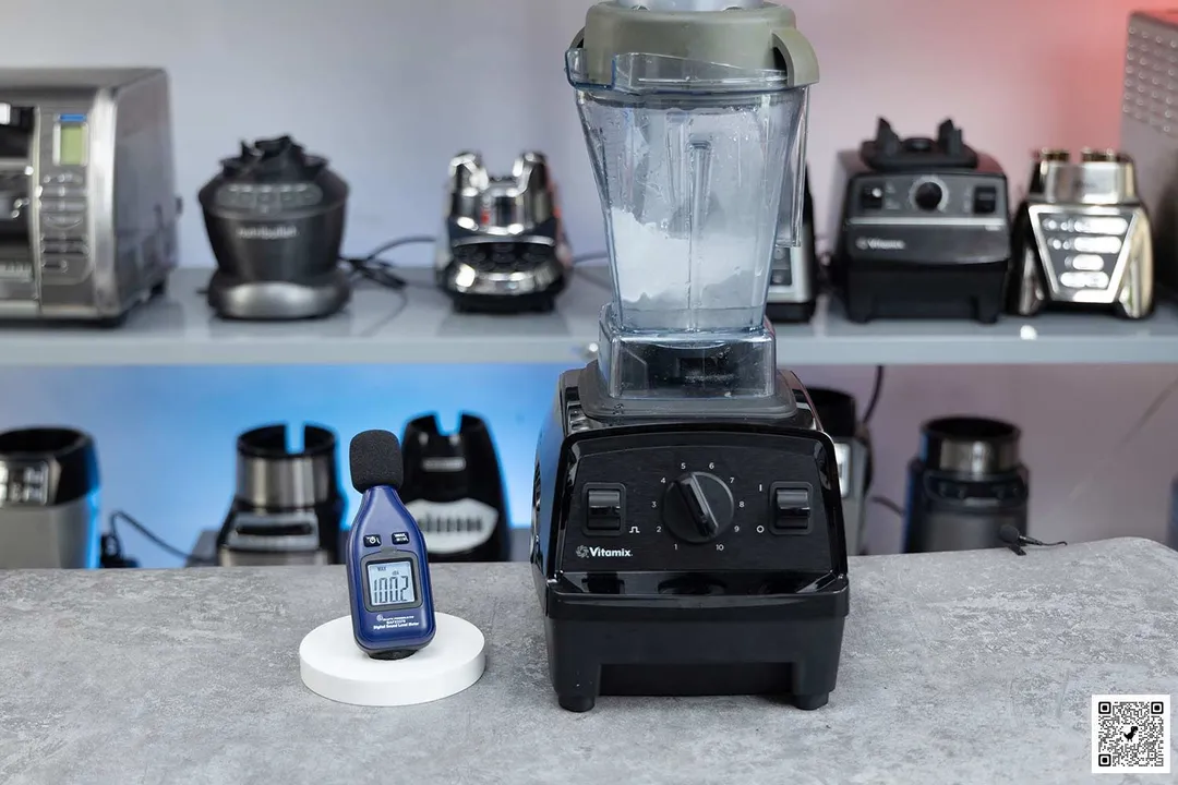 The Vitamix E310 blender is on a table, accompanied by a sound level meter displaying its noise level. In the background, other blenders that we tested sit on a nearby shelf.