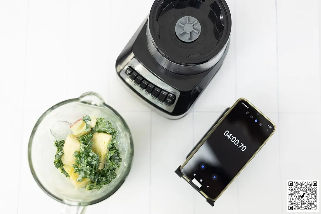 The Hamilton Beach Wave Crusher blender motor base and its container filled with apple, banana, and kale, stand on a table. Next to it, a smartphone showcases the blending time of 4 minutes.