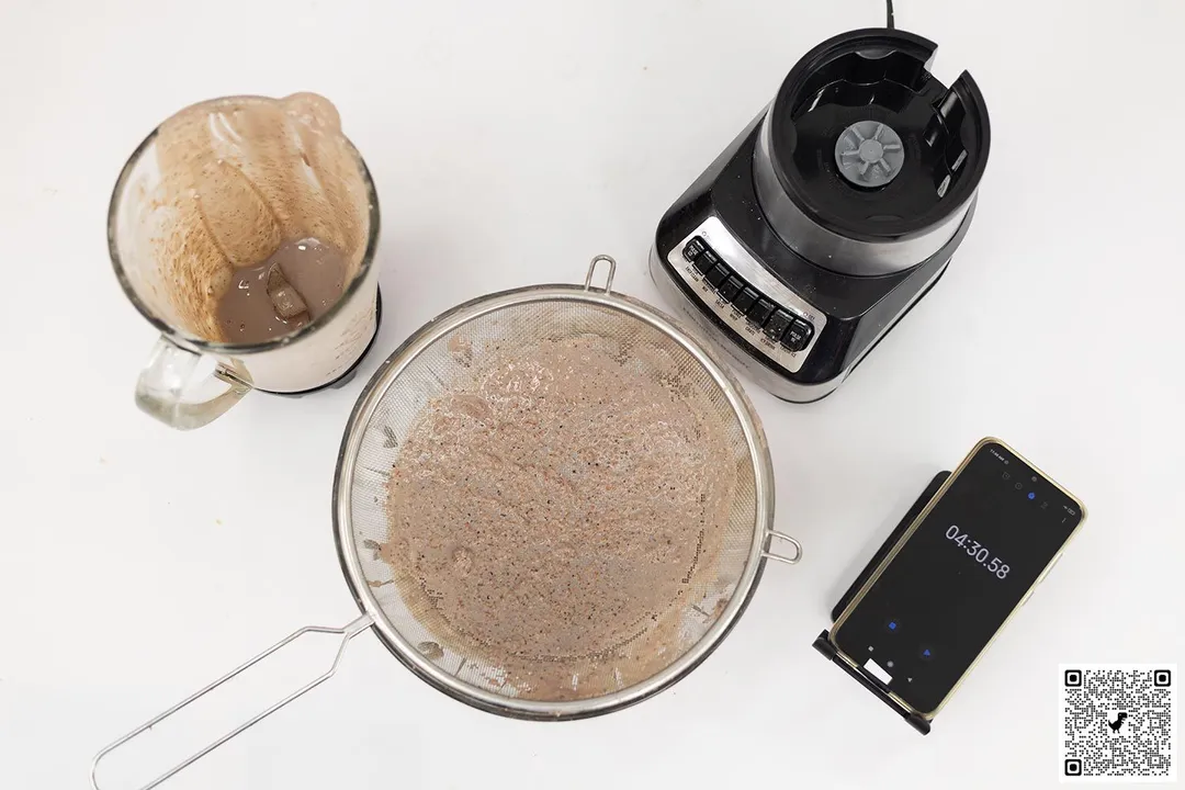 The Hamilton Beach Wave Crusher motor base stands beside the container. Next to it, a protein shake has been strained through a metal mesh strainer while a smartphone displays a blending time of 4 minutes 30 seconds.