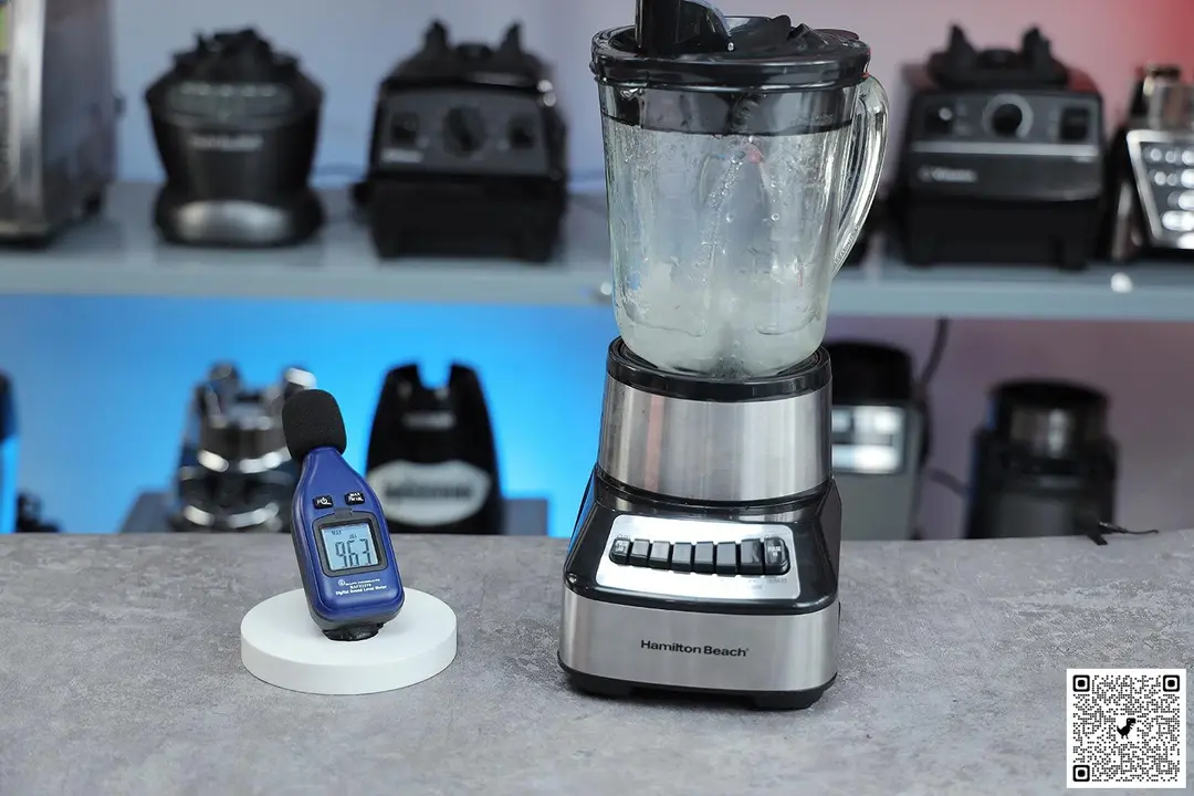 The hamilton beach wave crusher blender blender is on a table, accompanied by a sound level meter displaying its recorded noise level. In the background, other blenders that we tested sit on a nearby shelf.