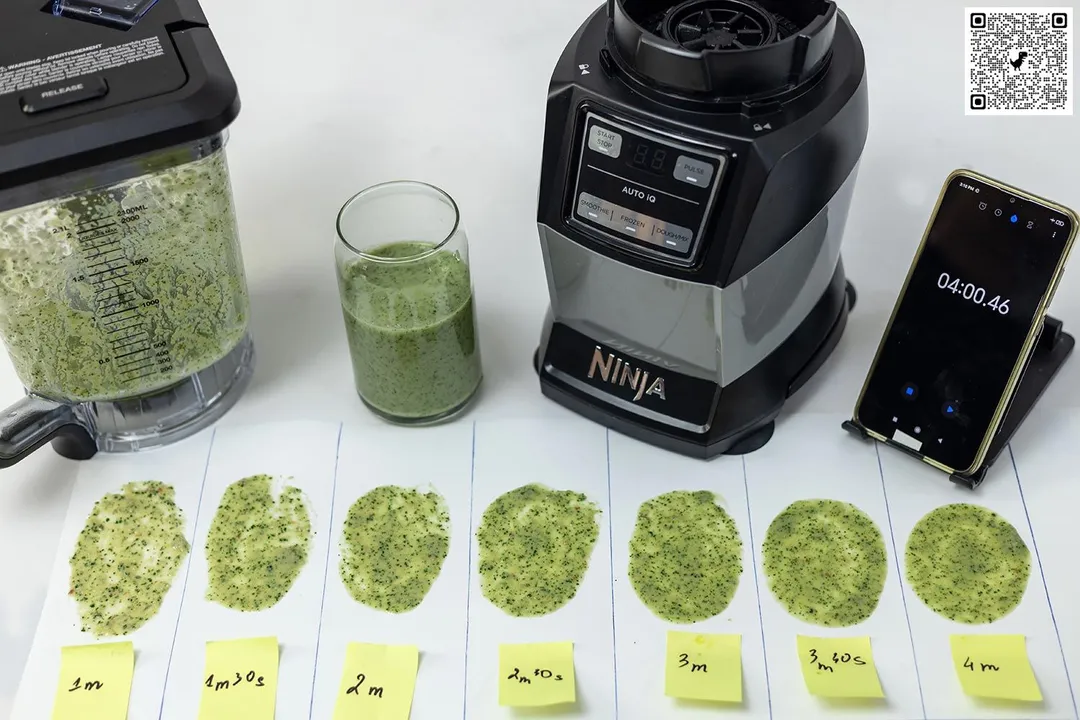 The AMZ493BRN blender’s motor base and its container stand on a table, accompanied by a glass filled with a green smoothie. Next to it, a white paper features three distinct smoothie samples while a smartphone showcases the blending time of 4 minutes.