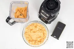 The Ninja AMZ493BRN motor base stands beside a glass bowl containing a portion of blended smoothie. Next to it, a smartphone displays a blending time of 4 minutes and 10 seconds.