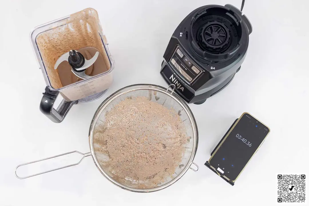 The Ninja AMZ493BRN blender motor base stands beside the container. Next to it, a protein shake has been strained through a metal mesh strainer while a smartphone displays a blending time of 3 minutes 40 seconds.