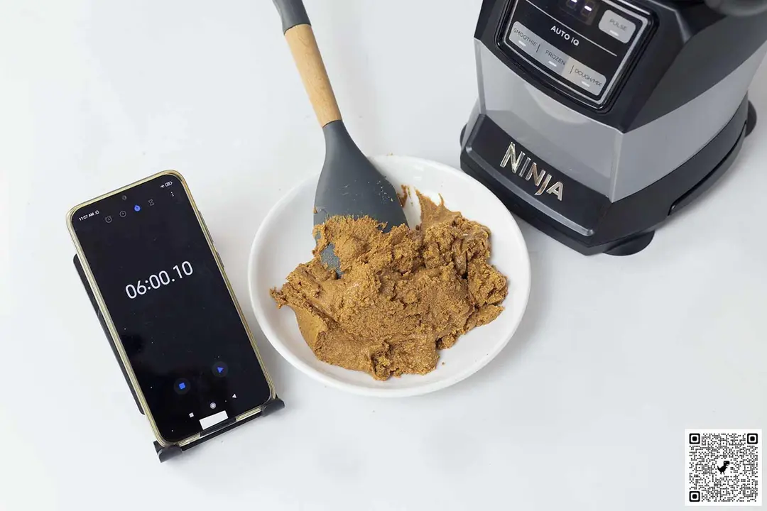 The Ninja AMZ493BRN is beside a white plate containing almond butter with a spatula, blend assembly, and a smartphone revealing a blending time of 6 minutes.