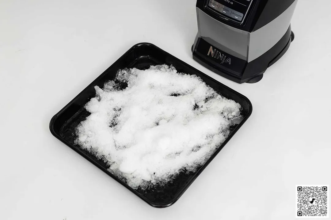 The Ninja AMZ493BRN blender is beside a black tray containing ice it crushed.