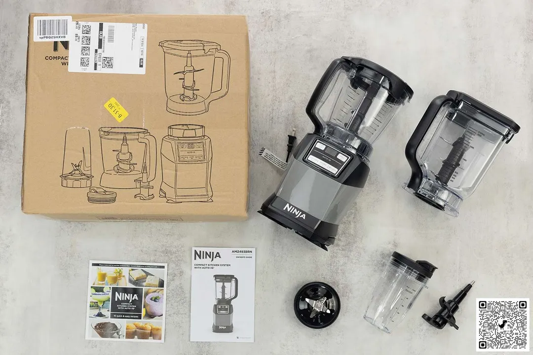A display of the Ninja Compact Kitchen System Blender and its accessories, including the 40-ounce processor bowl, dough blade assembly, pro extractor blades assembly, 18-ounce single serve cup, user manual, and carton box, on a table.