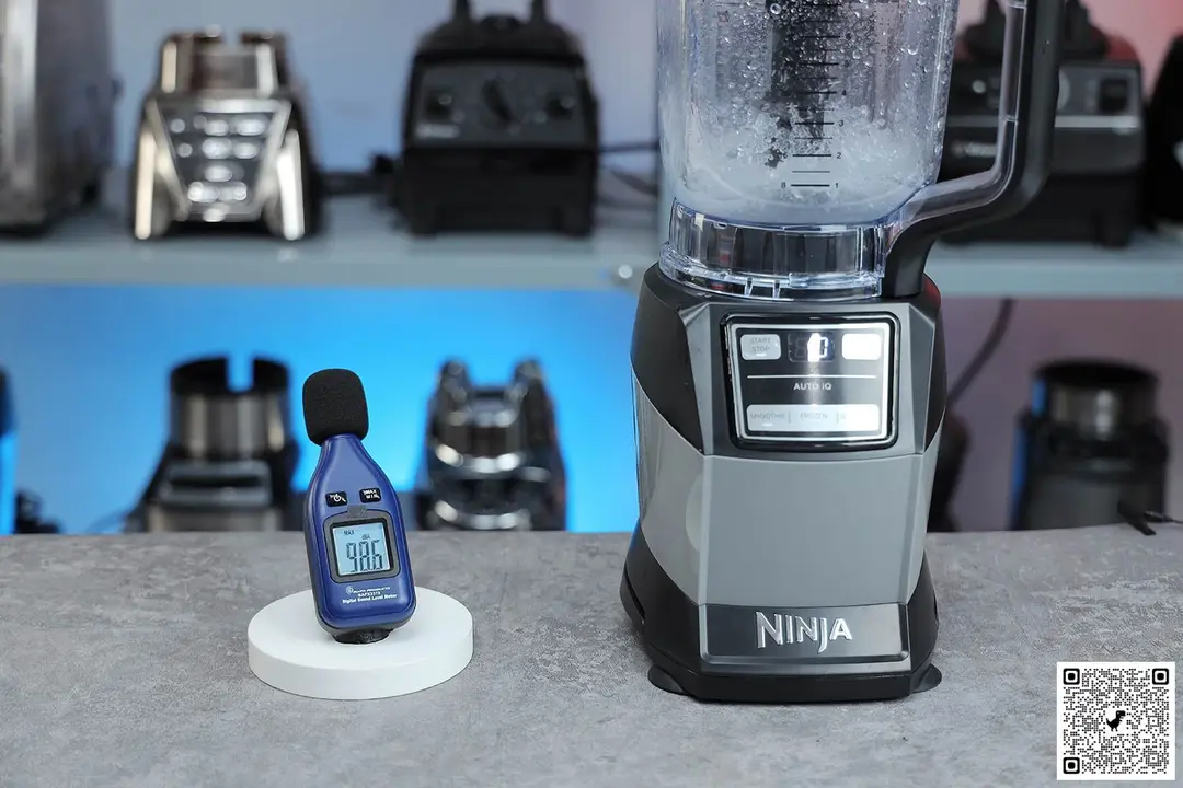 The Ninja Compact Kitchen System Blender is on a table, accompanied by a sound level meter displaying its recorded noise level. In the background, other blenders that we tested sit on a nearby shelf.