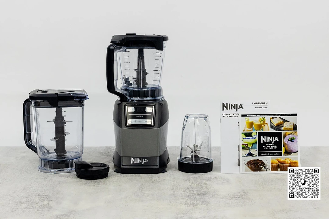 The Ninja Compact Kitchen System Blender and its accessories, including a processor bowl, single-serve blending cup, plastic lid, recipe booklet, and user manual, are on a table.