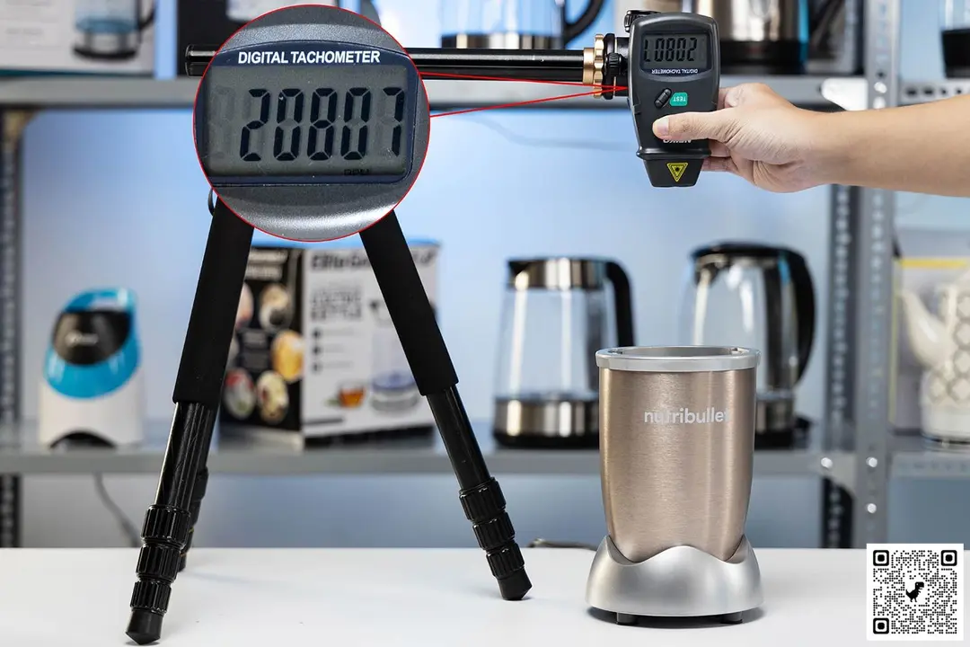 The NutriBullet Pro 900-Watt motor base on table with a hand pressing a digital tachometer on a tripod. Different blenders are visible in the background.