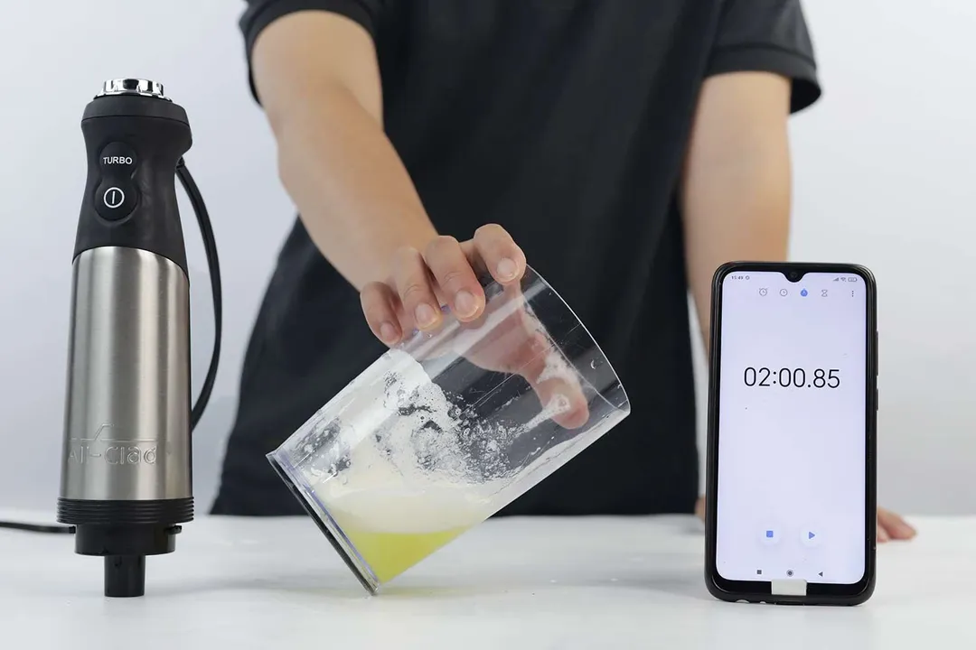 A person performs a stiffness test on whipped egg whites by tilting a beaker sideways, with an All-Clad immersion blender and a smartphone showing a stopwatch at 2 minutes and 0.85 seconds in the background.