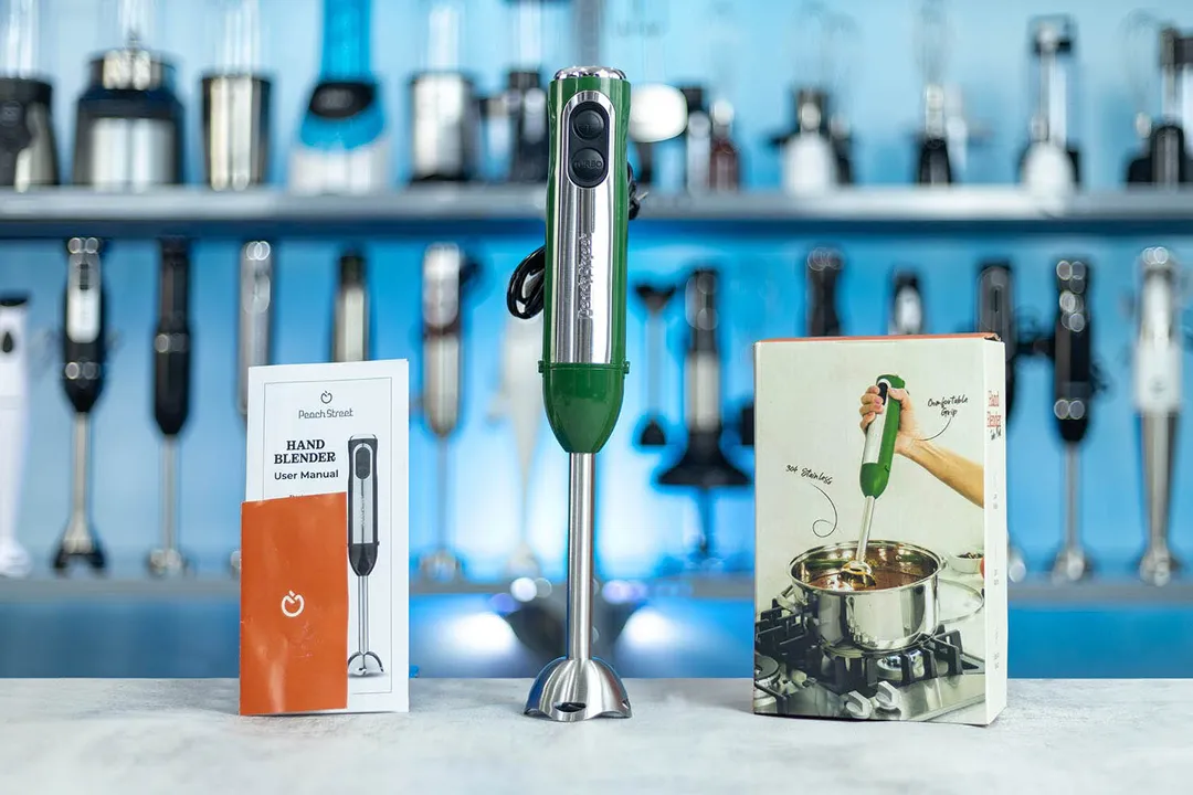 The Peach Street hand blender is displayed on a kitchen counter, accompanied by its user manual and a cookbook, with a backdrop of assorted kitchen appliances.