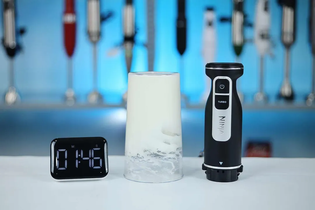 The Ninja CI101 Foodi immersion blender stands next to a glass of whipped egg whites forming stiff peaks, with a digital timer showing "1:46" in the foreground.