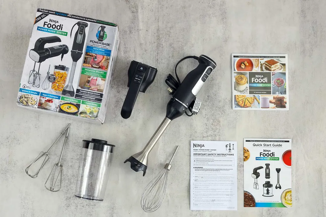The Ninja Foodi Power Mixer System displayed with its box, various attachments including a whisk, beater, and a blending cup, and instructional materials laid out on a gray countertop.