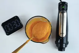Scooping a spoon of pureéd soup from a full batch in a plastic beaker made by the Braun MQ7035X immersion blender in 30 seconds.