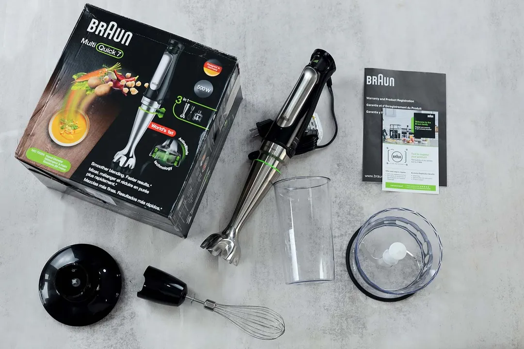 The Braun MQ7035X immersion blender unpacked beside its box, with accessories including a whisk and a chopper attachment, a blending beaker, and warranty and product registration paperwork.