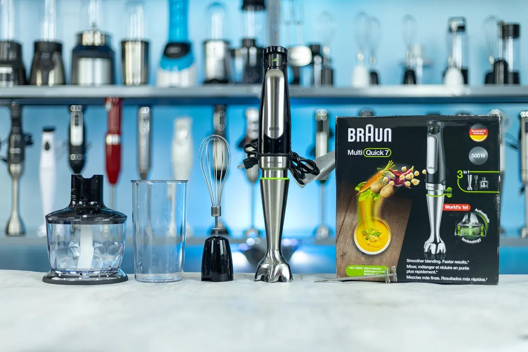 The Braun Multiquick 7 hand blender standing on a table with its accessories, including a plastic beaker, whisk attachment, food processor, and a paper carton box.