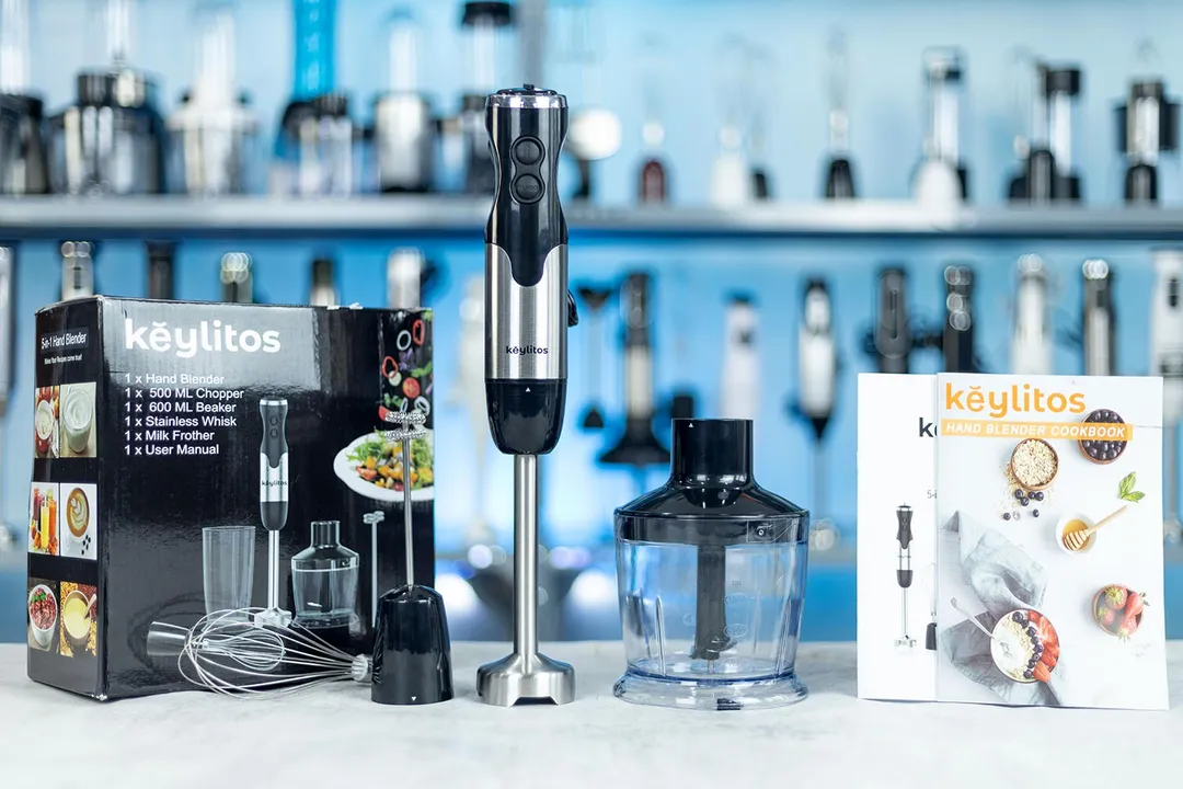 The Keylitos 5-in-1 hand blender set with its components and a cookbook on a kitchen counter, against a backdrop of shelves stocked with kitchen items.