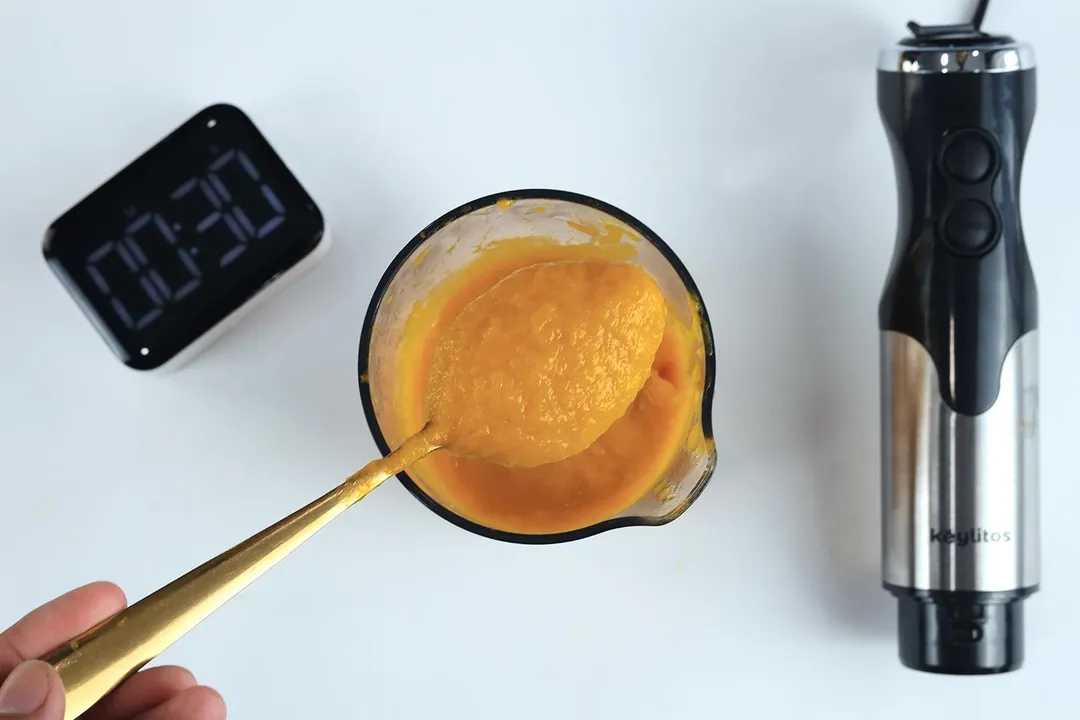 Scooping a spoon of pureéd soup from a full batch in a plastic beaker made by the Keylitos 5-in-1 immersion blender in 30 seconds.