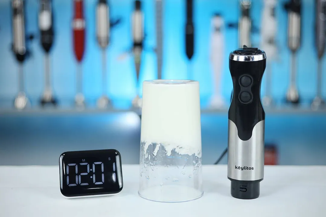 The Keylitos 5-in-1 immersion blender stands next to a glass of whipped egg whites forming stiff peaks, with a digital timer showing "2:01" in the foreground.
