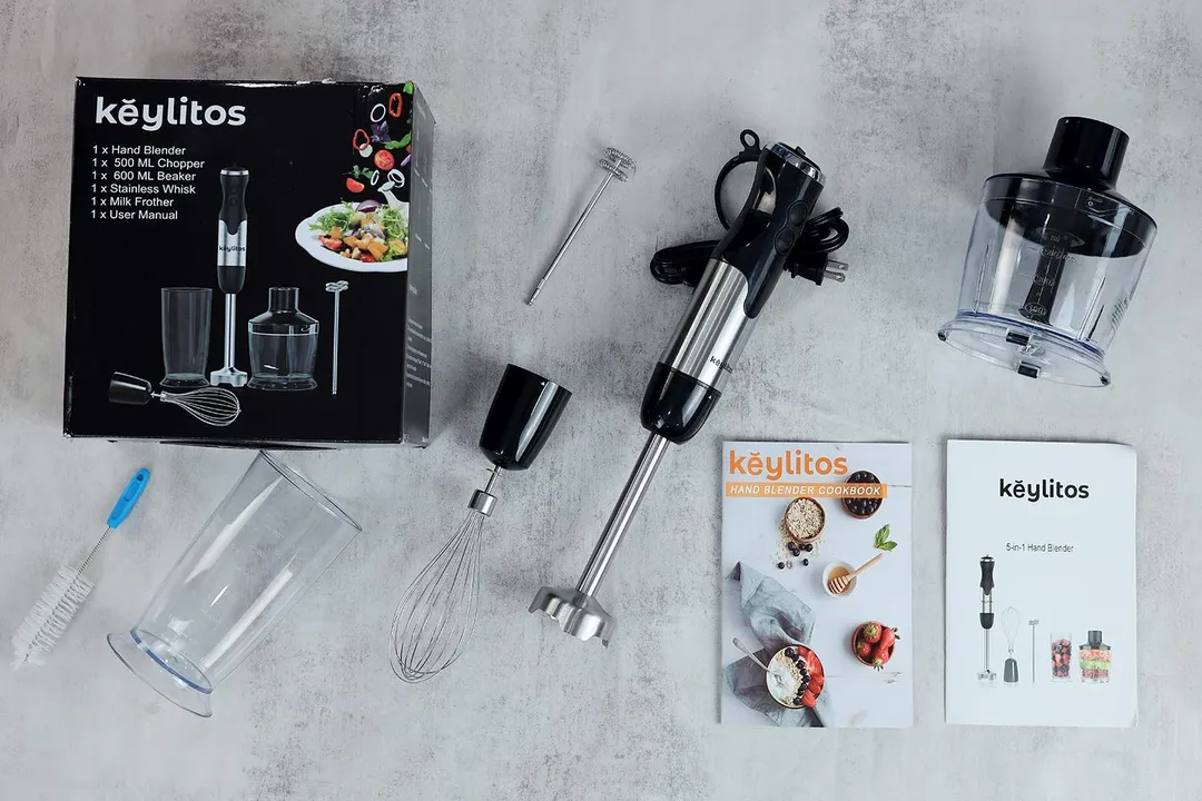The Keylitos 5-in-1 immersion blender lying on a table with its accessories, including a beaker, whisk, chopper, milk frother attachment, bottle brush, and user manual.